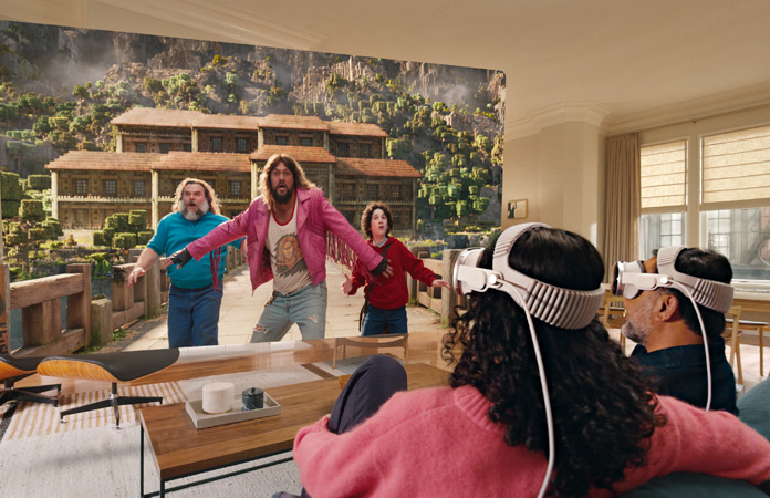 Two people both wearing Apple Vision Pro sit side by side on a couch watching: A Minecraft Movie, displayed on a screen stretching the across room